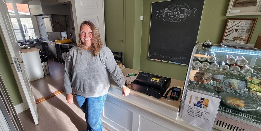 Fagarbeider Anette Lønnhaug i grå genser og lyse jeans står ved den plassbygde, hvite kafedisken med pengekasse, matskap og korktavle på den grønne veggen bak disken. Hvit dobbeldør til venstre inn til kjøkkenet. Foto.