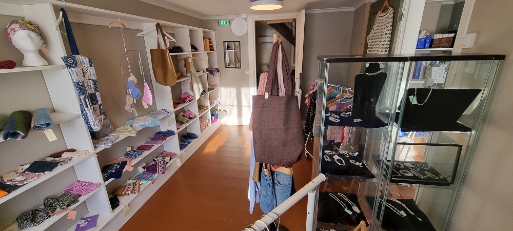 Butikk i første etasje med ting som brukerne har produsert, for eksempel strikketøy og smykker. Varene ligger på hyller i hvit reol, i et glassmonter og på en stumtjener. Foto.