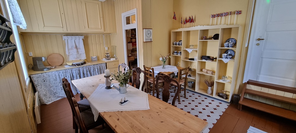 Gult rom, innredet som en gammel landhandel, gardinbrisk, hyller med antikke ting, avlangt bord med stoler og skafferi. Høyt under taket, trapp opp til hvit dør, brunt og hvit/sort rutete gulv. Foto.