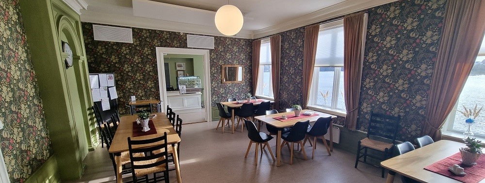 Kafe med blomstret tapet, grønnmalt klokkevegg, fem bord med stoler, kuppellampe midt i det høye rommet. Brune gardiner på hver side av de tre store vinduene ut mot sjøen. Dør inn til disk og kjøkken. Foto.