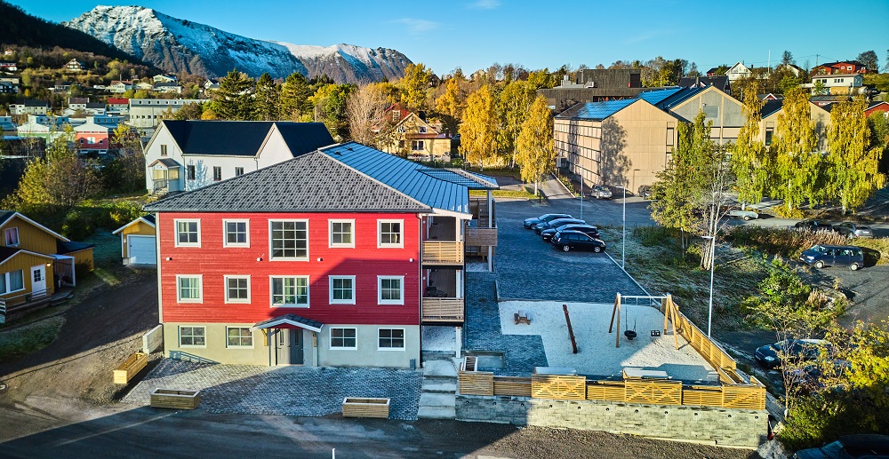 Dronefoto forfra, lekeplass til høyre, bebyggelse og trær omkring, snødekte fjell i bakgrunnen. Foto.