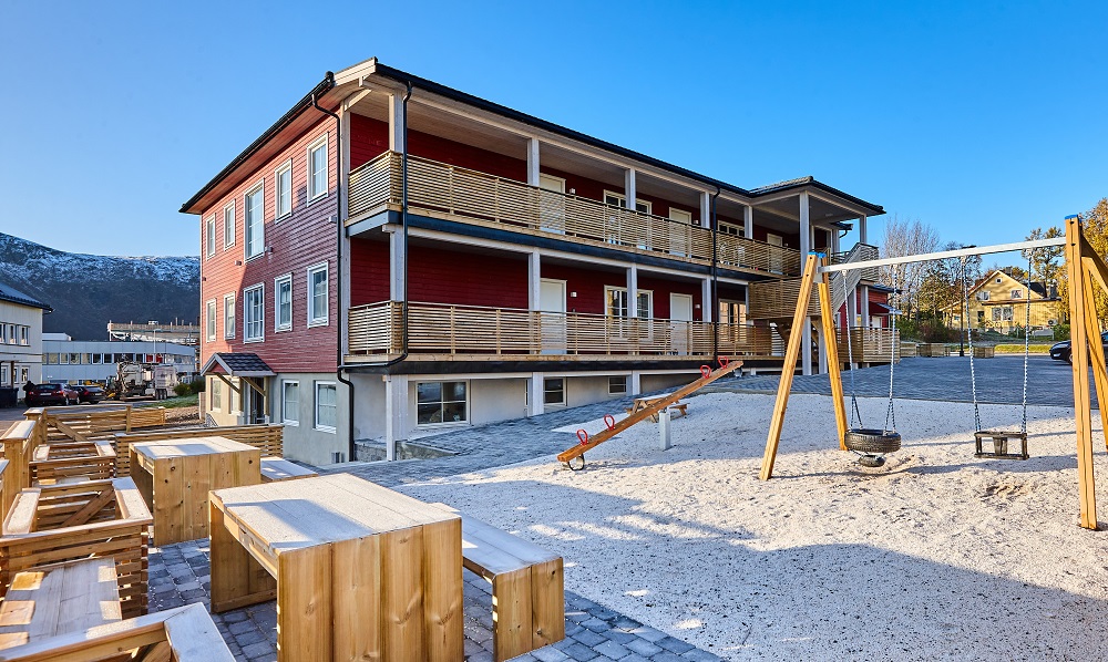 Lekeplass rett ved siden av leilighetsbygget med benker og bord av tre. Blå himmel. Foto. 