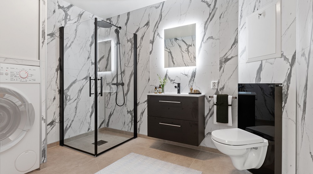 Romslig bad, lyse baderomsplater med grått mønster på veggene, belegg på gulv. Dusjhjørne, servant med to skuffer, lysspeil og vegghengt klosett. Foto.