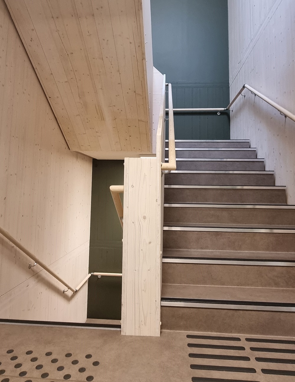 Trappeoppgang med lyse massivtre vegger, rekkverk og markeringer i trinn og gulv for svaksynte. Foto. 