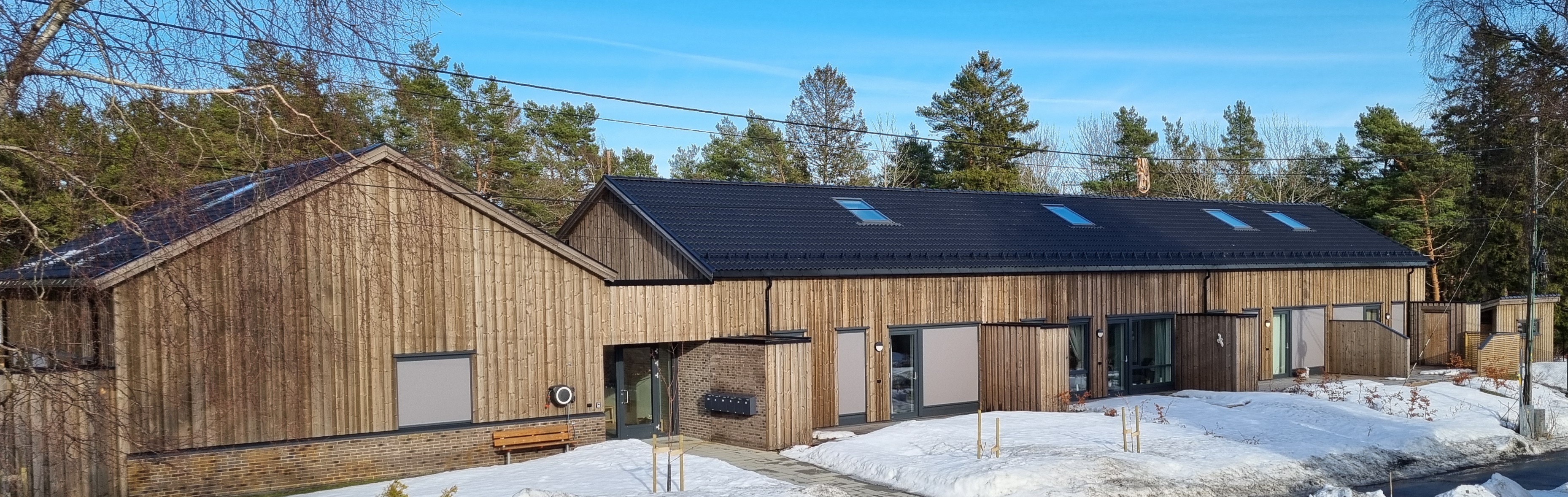 Fasade langside i én etasje med trebrune vegger, terrasse med skillevegger, sorte tak med takvinduer. Snødekt bakke mellom hagen og veien. Foto. 