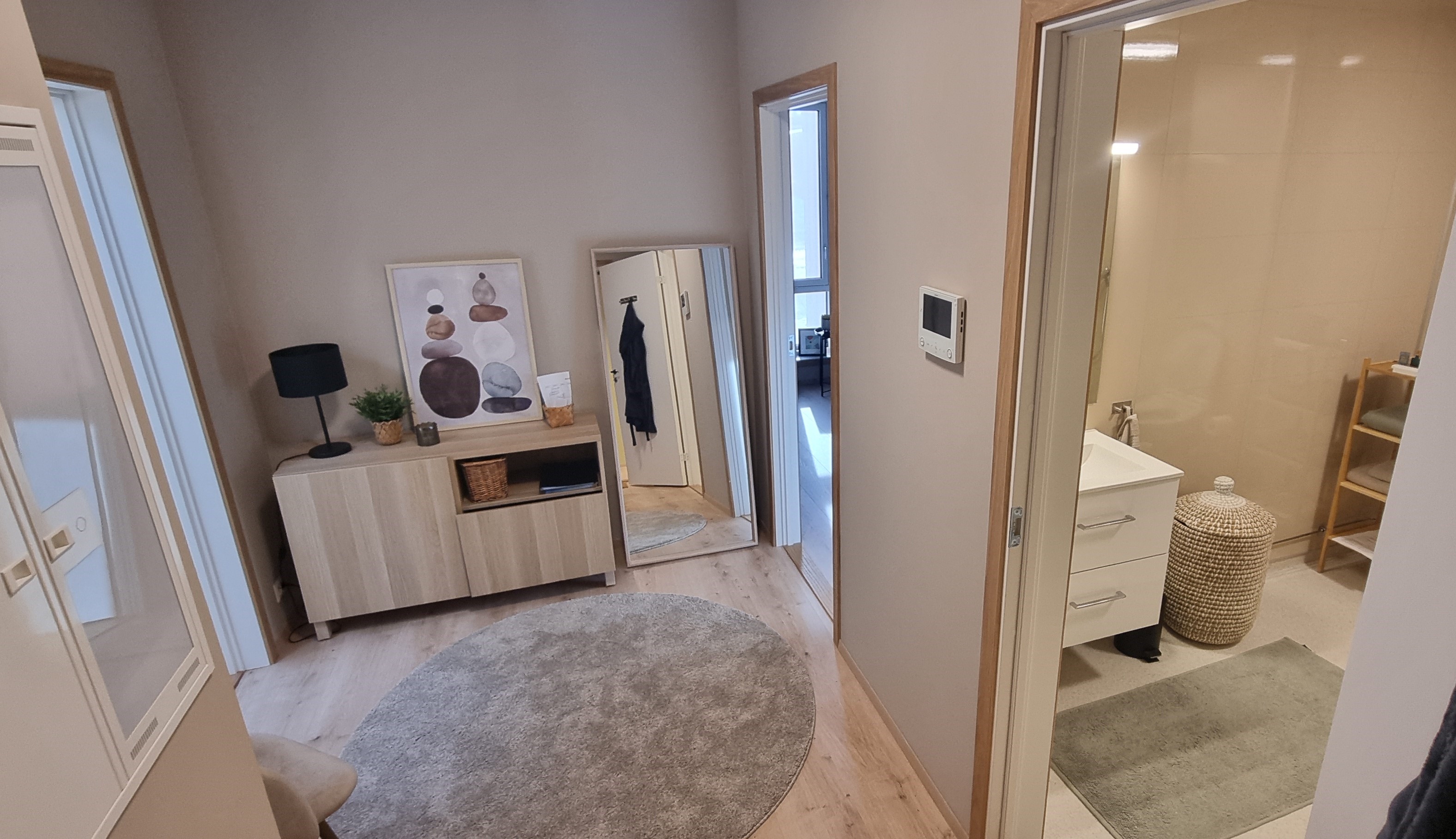 Mellomgang i hver leilighet med bad til høyre, soverom innerst til høyre, stur-kjøkken innerst til venstre. Lyse og varme farger, rundt teppe, speil og kommode på gulvet. Foto. 