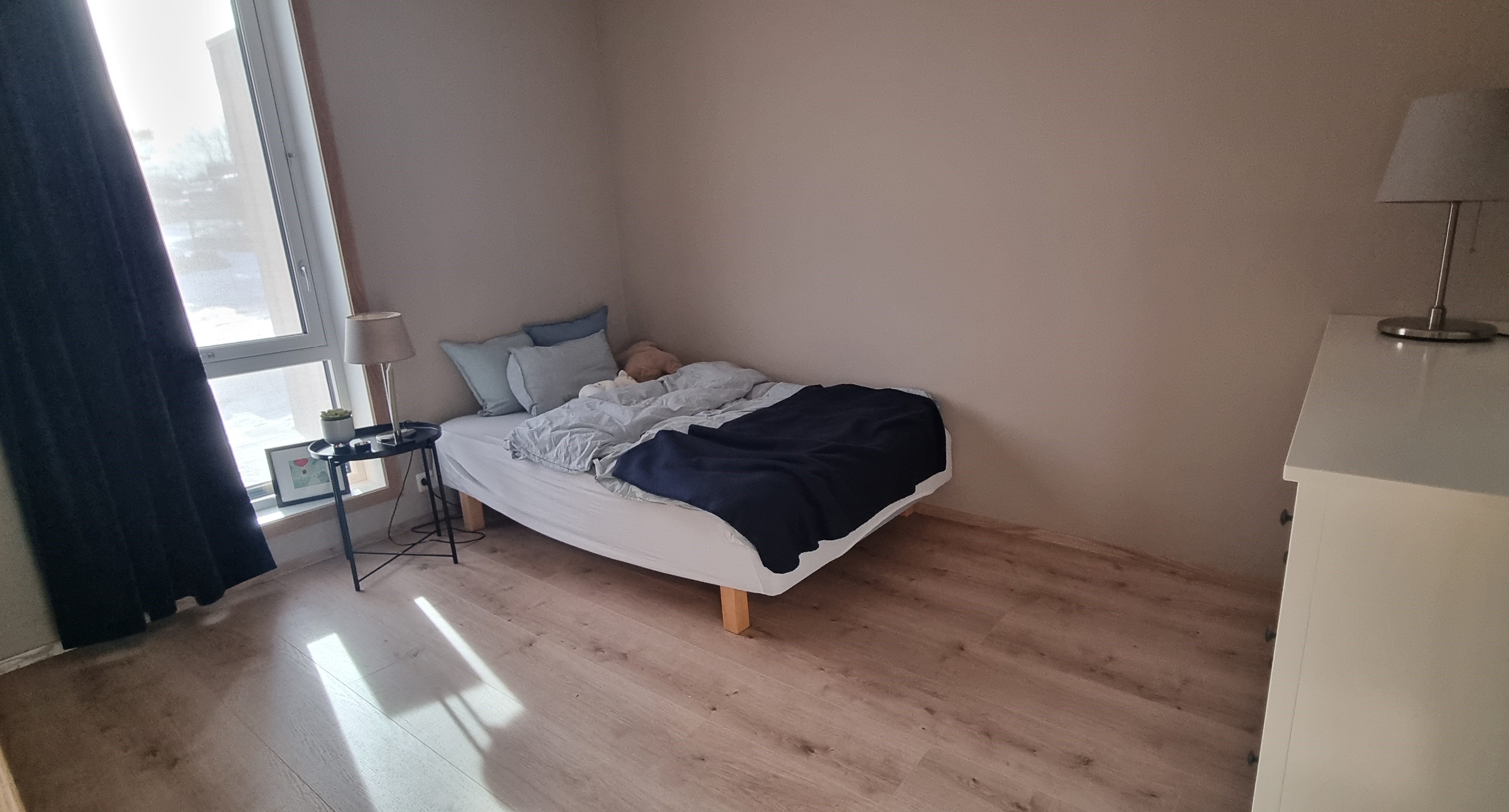 Soverom med vindu fra gulv til tak og blendegardin. Oppredd seng, kommode med lampe- Lyse og varme farger. Foto.