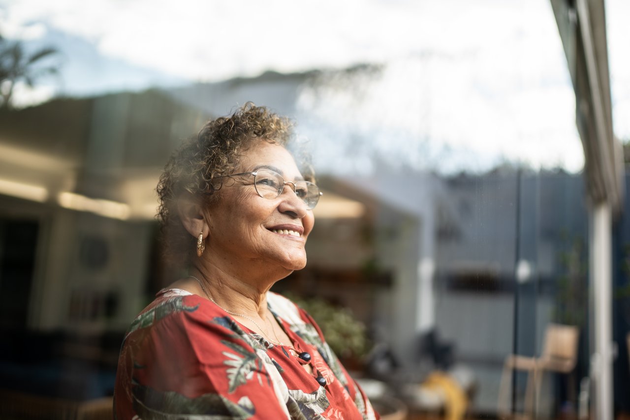 En eldre kvinner ser smilende ut av vinduet i boligen sin. Foto.
