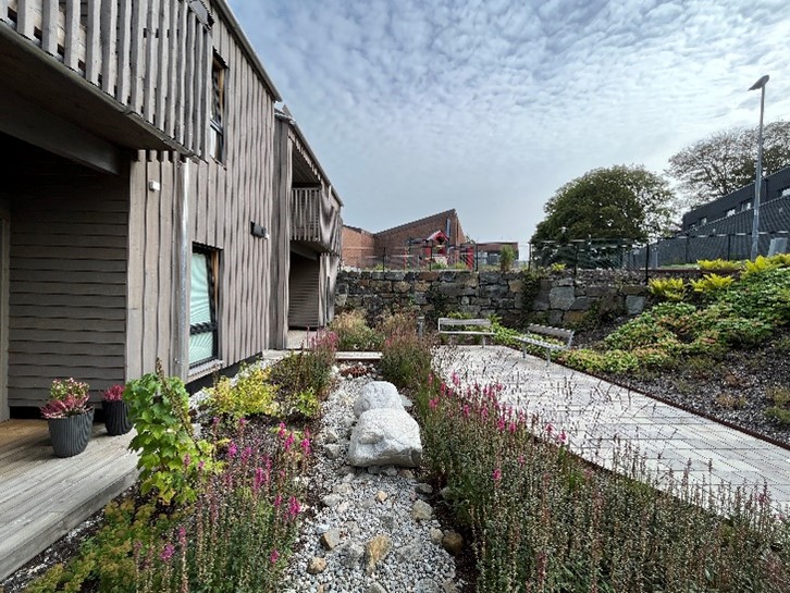 Vi ser huset i to etasjer med grå trefasade. Terrasser i første etasje og balkonger i andre etasje. Blomster og sitteplasser. Foto.