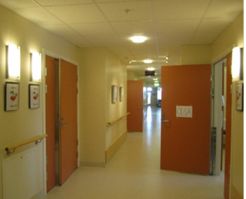 Korridor med dører på begge sider og veggen ved dørene er trukket litt inn. To av dørene er åpne og viser at dørbladet slår ut i korridoren. Foto.