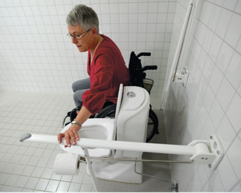 Dame i rullestol ved siden av toalett. Holder hånd på armstøtte på motsatt side av toalettet. Foto.
